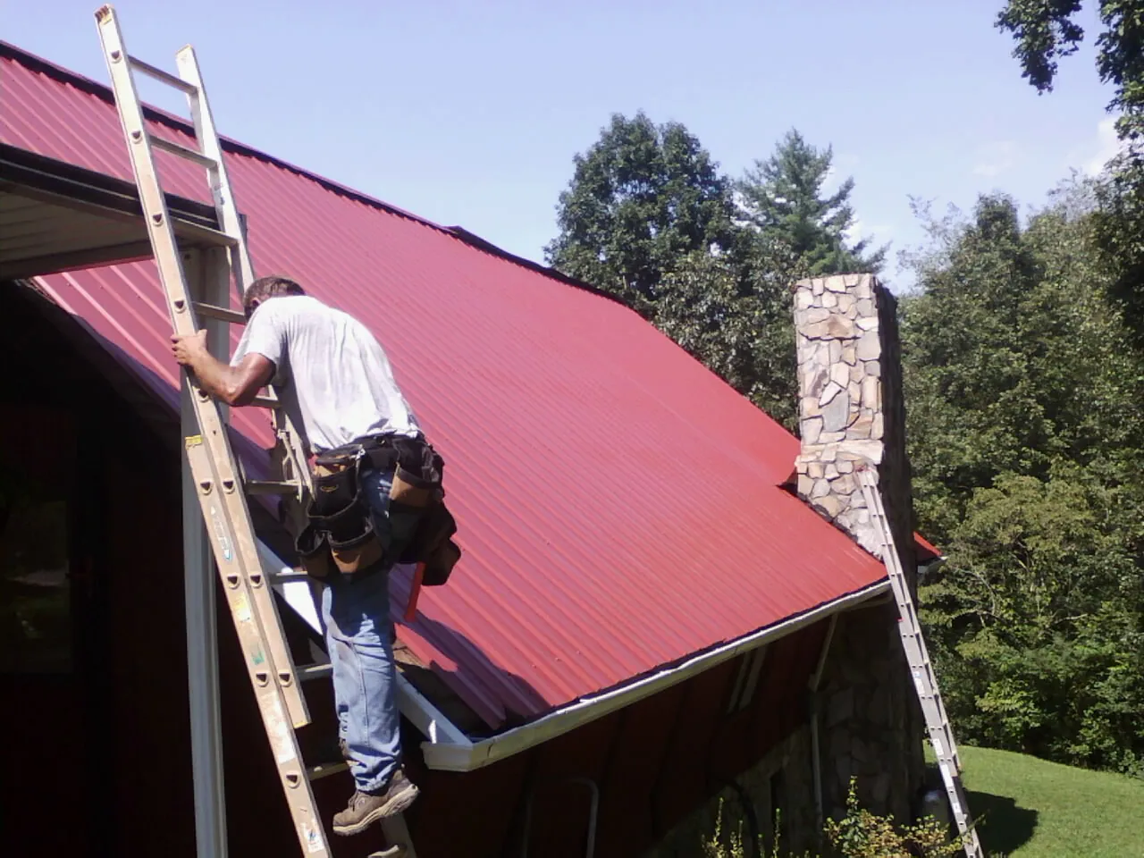 Roofer on a ladder beside a red metal roof and stone chimney — Soffit & Fascia Repair by Roofing Brothers in Hebron